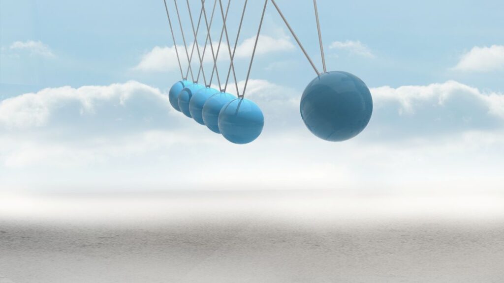 A blue Newton's cradle suspended against a soft sky with clouds, with one ball gently pulled back before it begins to swing.