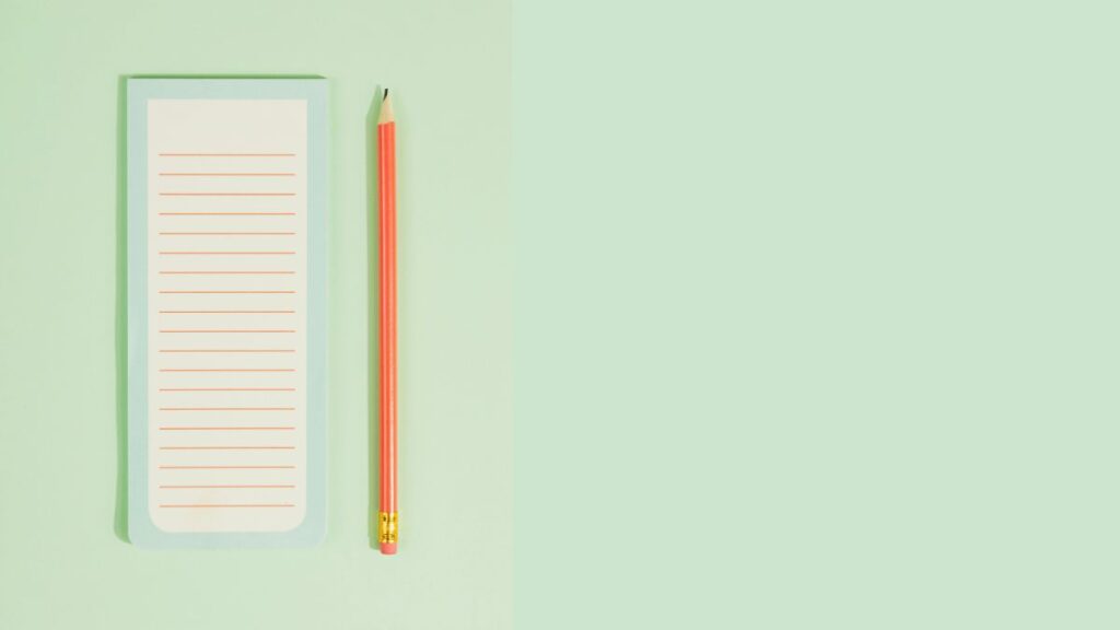 A small lined notepad and a coral pencil rest on a soft pale green background with plenty of open space to the right, creating a simple, uncluttered scene.