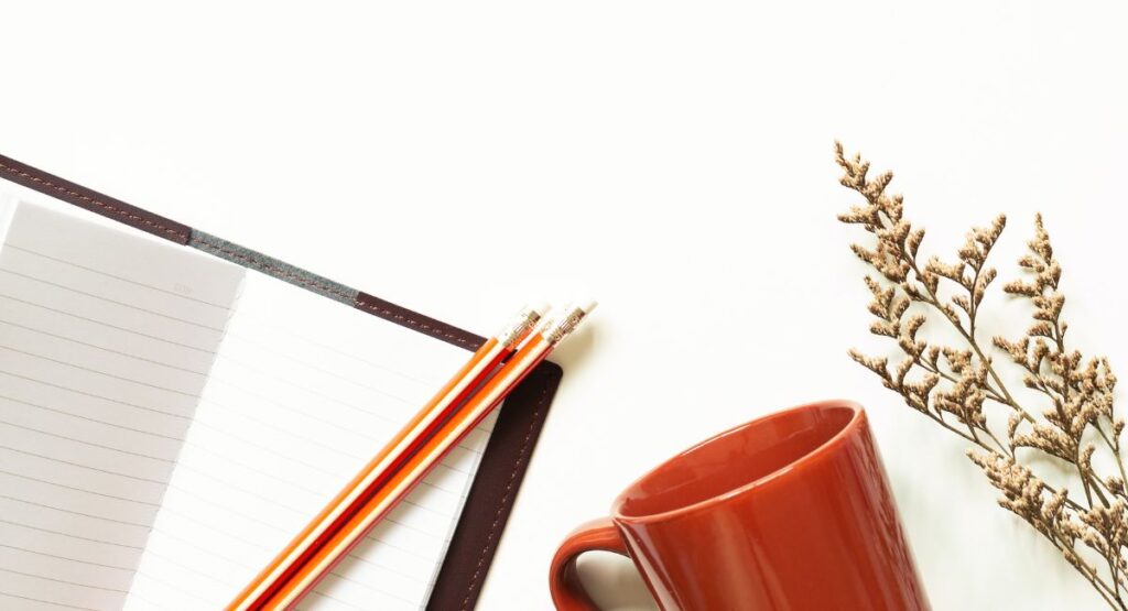 Open notebook with pencils, a terracotta mug and dried flowers on a white desk, representing a quiet space for reflection and gentle momentum.