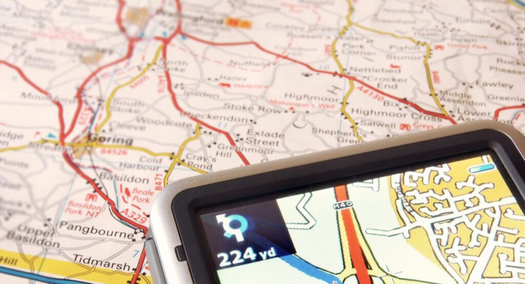 A handheld TomTom sat nav resting on top of an open paper road map, its screen showing a blue turn arrow and '224 yd' to the next junction.