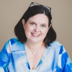 Jen Waller smiling broadly at the camera, glasses pushed up on her head. She has shoulder-length brown hair and wears a blue and white patterned top, a small pearl necklace, against a warm beige background.