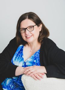 Jen Waller smiling and looking off to the side, resting her folded arms on the back of a cream, textured armchair. She wears glasses, a blue floral top, a black cardigan, and a small pearl necklace, against a plain light grey background.