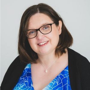 Headshot of Jen Waller, smiling warmly at the camera. She has shoulder-length brown hair and glasses, and wears a blue floral top with a black cardigan and a small pendant necklace, against a plain light grey background.