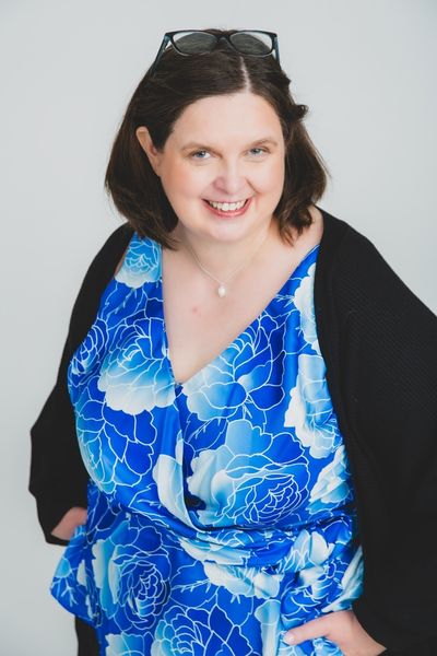 Jen Waller smiling warmly at the camera, glasses pushed up into her hair, wearing a blue floral top and black cardigan against a plain light grey background.