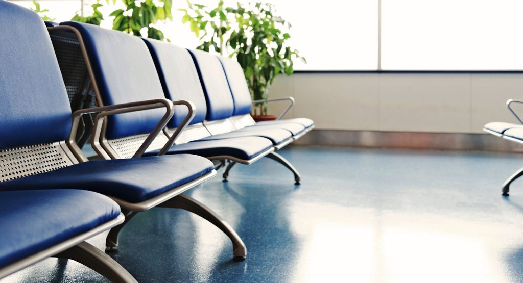 Row of empty blue waiting-room chairs beside a large sunlit window, with a green potted plant and a reflective floor.