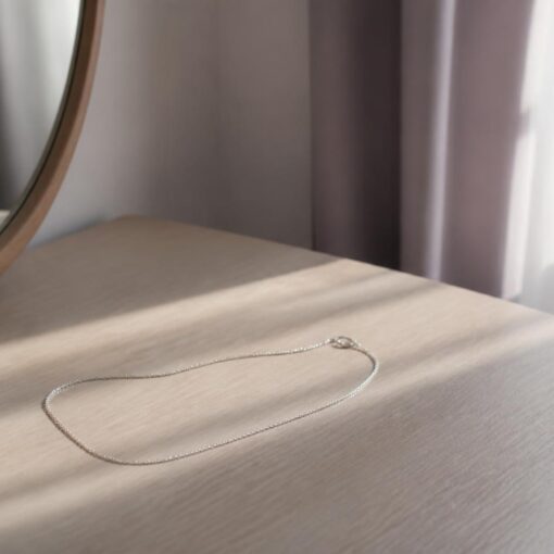 A fine silver chain necklace resting on a pale wooden dressing table beside a round mirror and a small ceramic dish, with soft sunlight falling through sheer curtains.