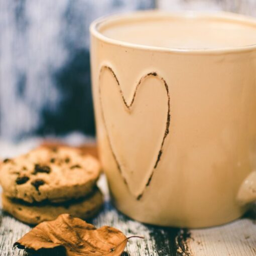 A cream-coloured mug with a heart on it, filled with a warm drink, beside two chocolate chip biscuits and a few autumn leaves on a weathered wooden table.