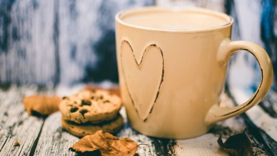 A cream-coloured mug with a heart on it, filled with a warm drink, beside two chocolate chip biscuits and a few autumn leaves on a weathered wooden table.