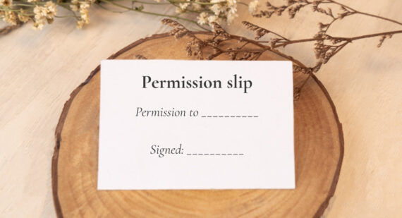 A white card resting on a slice of wood, with dried flowers above. Text on the card reads: Permission slip. Permission to, followed by a blank line. Signed, followed by a blank line.