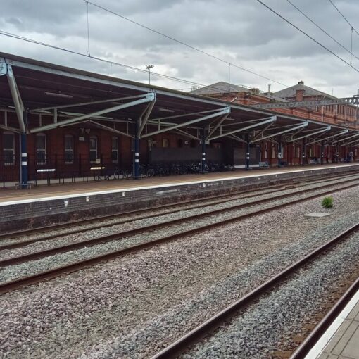 A view along the platform at Rugby station on a cloudy day, with empty railway tracks, a long red-brick building with a metal-framed canopy, and bicycles parked beneath it.
