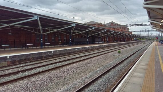 A view along the platform at Rugby station on a cloudy day, with empty railway tracks, a long red-brick building with a metal-framed canopy, and bicycles parked beneath it.