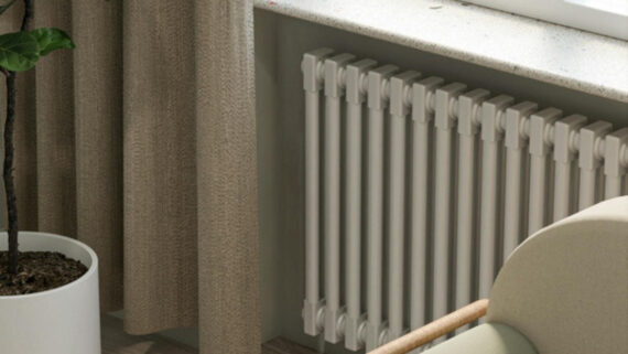 A cream column radiator beneath a window sill, with a leafy plant and a soft curtain beside it